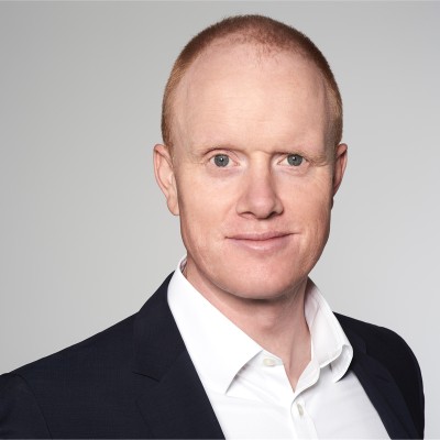 A professional headshot of a man with short red hair and a slight smile, wearing a dark suit and white shirt against a light gray background.