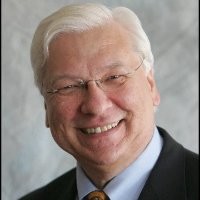 A smiling older man with white hair wearing glasses and a suit, set against a neutral background.